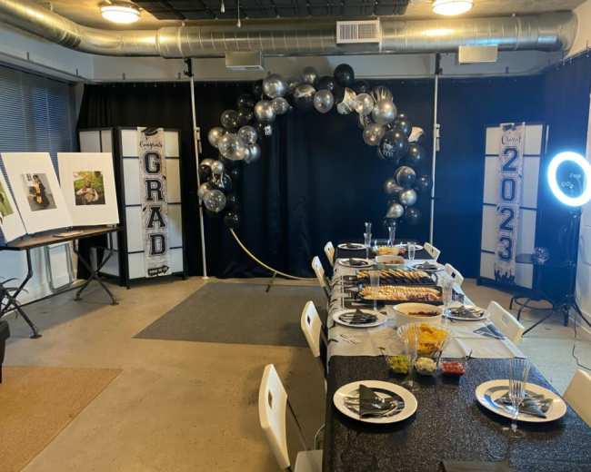 The image shows a decorated event space with a long table set for a celebration, featuring a black and silver balloon arch in the background and graduation-themed displays.