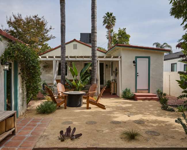 The image shows a small courtyard with wooden chairs arranged around a fire pit, framed by palm trees and surrounded by greenery.