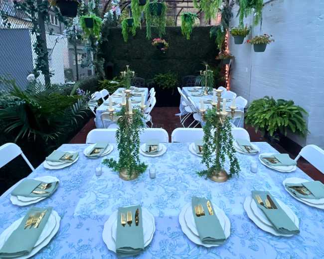 A decorated outdoor dining area features a long table set with gold utensils and green napkins, surrounded by hanging plants and fairy lights.