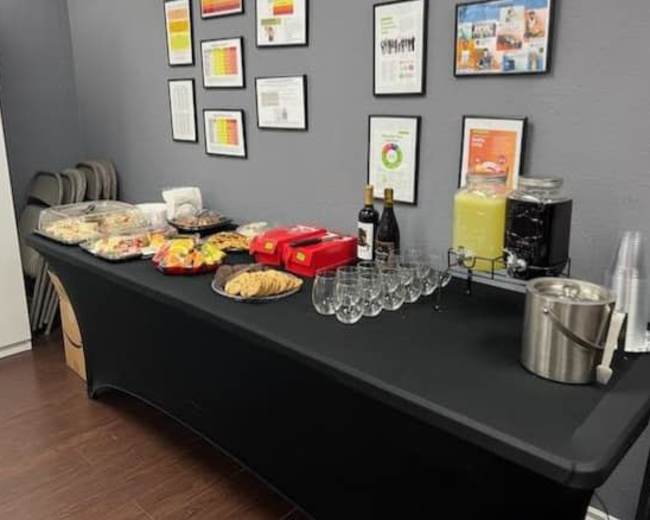 A table is set up against a wall, displaying a variety of food items, drinks, and glassware, with several framed documents on the wall behind it.
