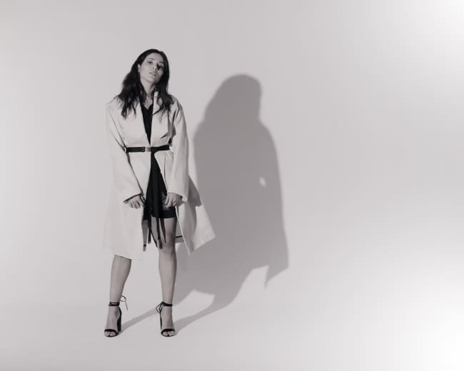 A person stands in front of a plain white background, wearing a light-colored coat and high-heeled sandals.