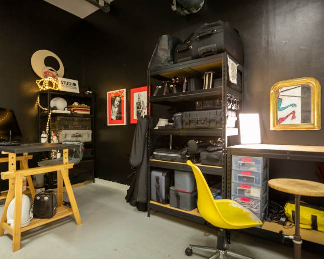 The image shows a compact workspace with black walls featuring shelves of equipment, a wooden desk with a monitor, and a yellow chair.