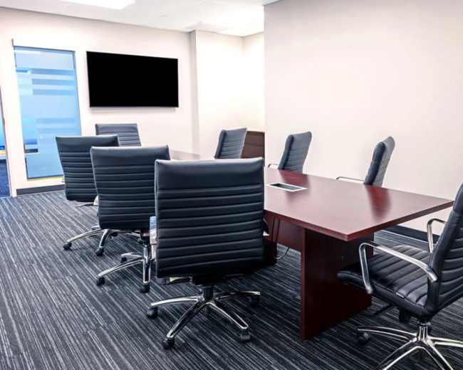A modern conference room features a long wooden table surrounded by six black leather chairs, with a large TV screen mounted on the wall.