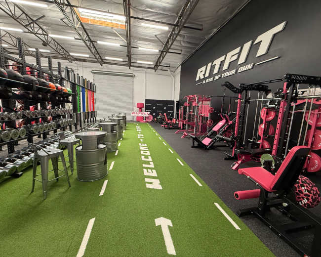 The image shows a fitness gym featuring various exercise equipment, weights, and a turf pathway with a welcome message.