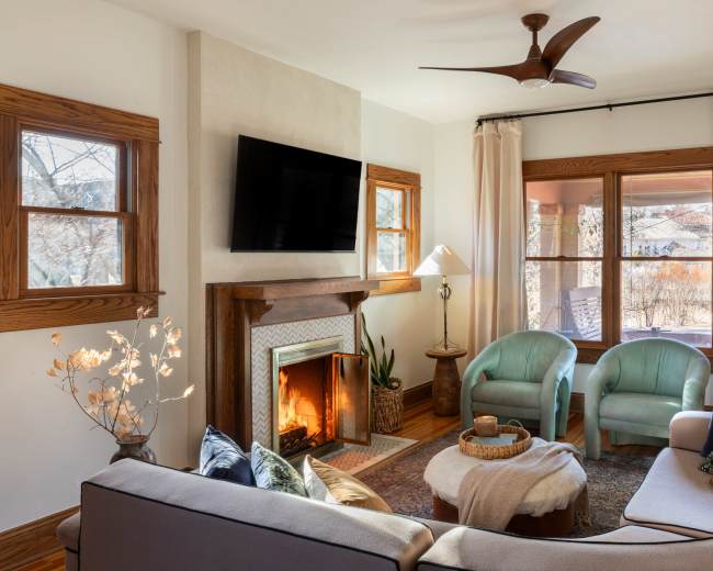 A cozy living room featuring a fireplace, two light blue chairs, a round coffee table, and large windows with natural light.