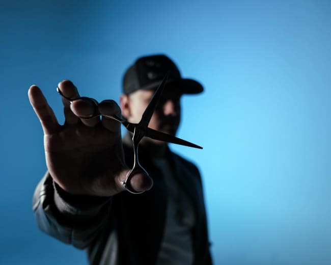 A person in a dark jacket and cap is holding a pair of scissors in front of them against a blue backdrop.