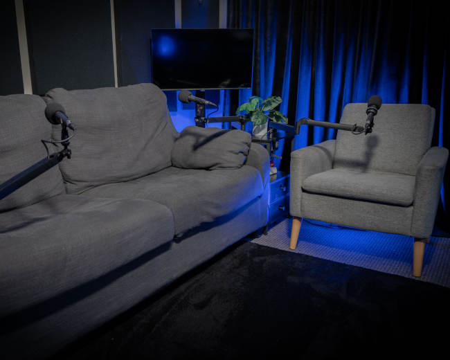 The image shows a cozy recording studio setup with a gray couch and an armchair, both equipped with microphone stands, and a television in the background.