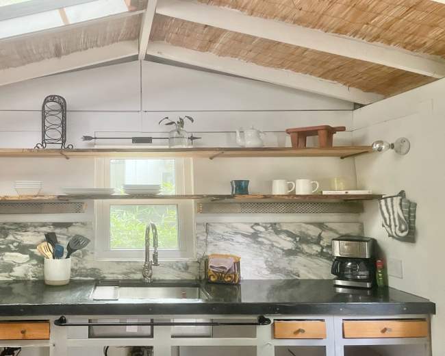 The image shows a kitchen with open shelves displaying dishware and plants, a black countertop, and a window allowing natural light to enter.