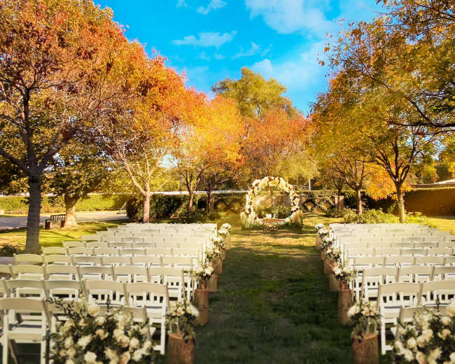 A wedding ceremony setup features rows of white chairs arranged on a grassy area surrounded by trees with autumn foliage.