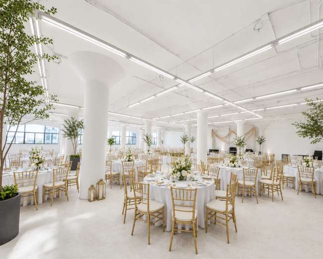The image shows a spacious banquet hall set for a formal event, featuring round tables with white tablecloths and gold chairs, adorned with floral centerpieces and greenery.