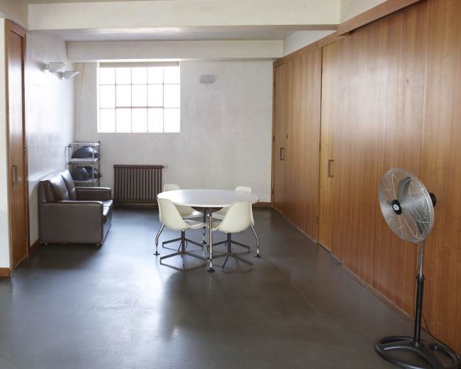 A minimalist room with a desk, television, brown sofa, round white dining table, and a standing fan.