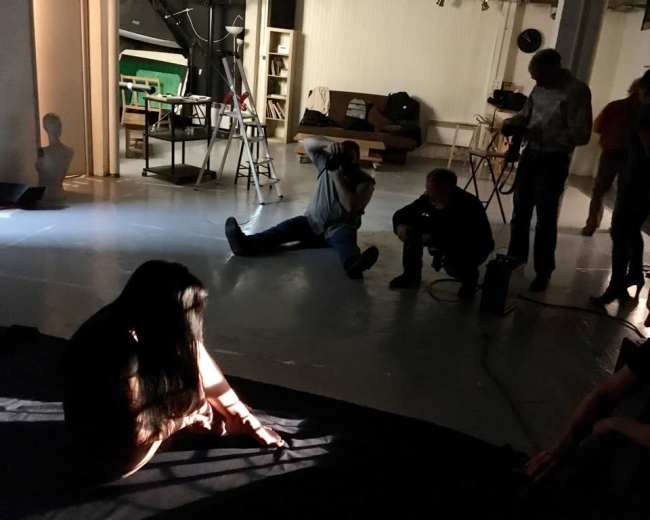 A group of people is gathered in a studio, with one person sitting on the floor and others engaged in activities around them.