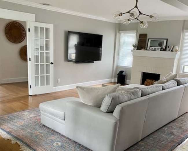 The image depicts a bright living room featuring a light-colored sofa, a large area rug, a television mounted on the wall, and a fireplace.
