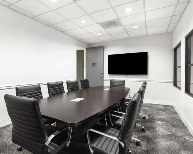 A modern conference room features a large table surrounded by black chairs, with a mounted screen on the wall and windows allowing natural light.