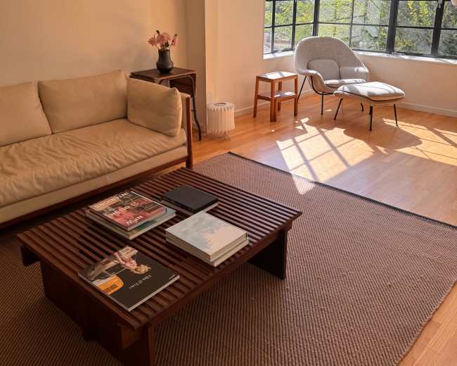 The image shows a bright, modern living room with a beige sofa, a wooden coffee table stacked with magazines, and a large window allowing natural light to enter the space.