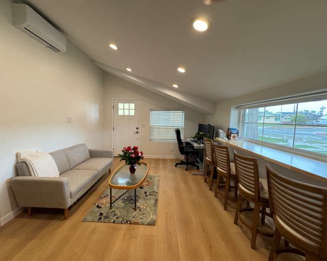 The image shows a modern living space with a gray sofa, a glass coffee table, and a long dining area with multiple wooden chairs, illuminated by ceiling lights.