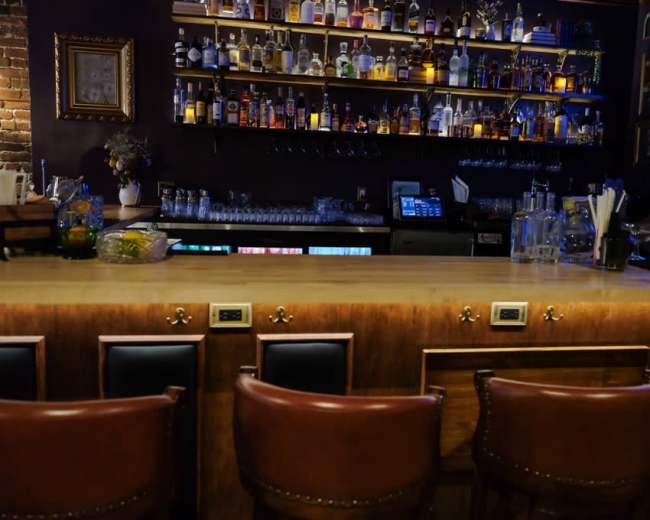 The image shows a dimly lit bar with a wooden counter and shelves lined with various bottles of liquor.