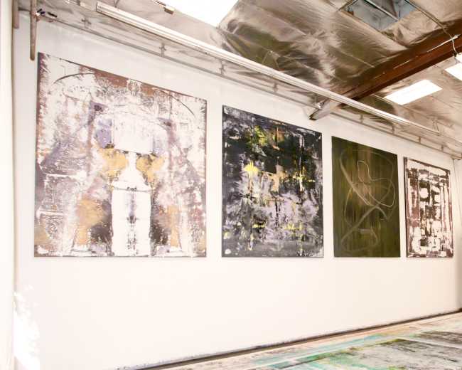 The image shows a gallery space with four large abstract paintings displayed on a white wall, alongside a patterned floor.