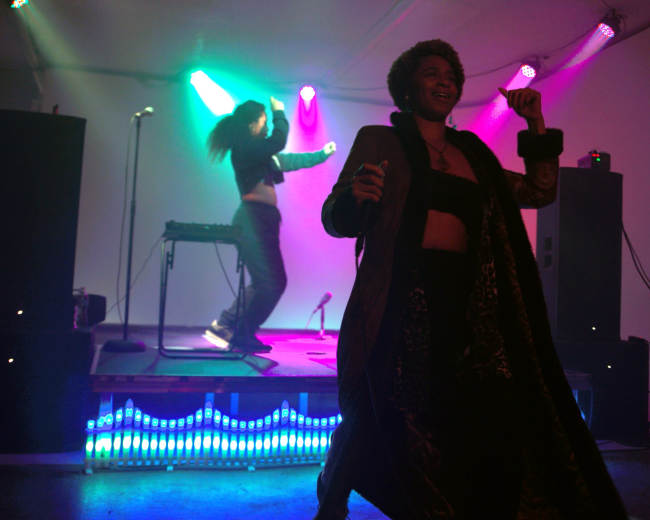 A woman in a long coat dances in front of a stage while another performer moves behind her under colorful lights.