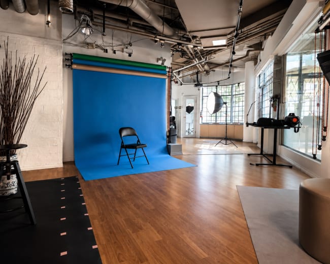 The image shows a photography studio featuring a blue backdrop, a black chair, and various lighting equipment arranged throughout the space.