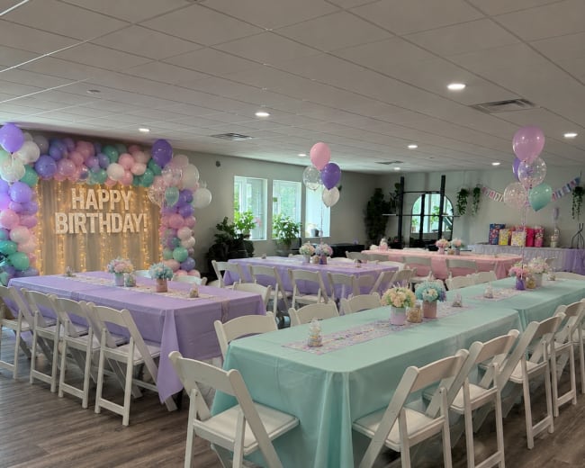 The image shows a decorated party space with tables set in alternating shades of lavender and mint green, surrounded by balloons and a "HAPPY BIRTHDAY" sign on the wall.
