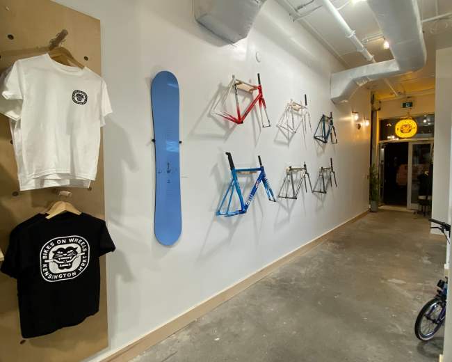 The image shows a hallway with a clothing display on the left and various bicycle frames mounted on the wall to the right, leading to an entrance at the far end.
