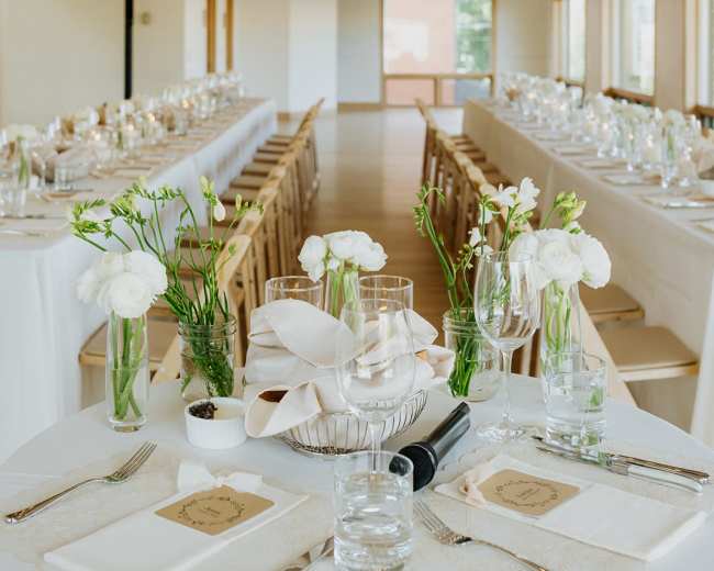 A beautifully arranged banquet hall features long tables with white linens, centerpieces of flowers and candles, surrounded by decorative greenery and soft lighting.