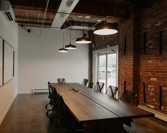 A large conference room features a wooden table surrounded by black mesh chairs, with whiteboards on the wall and industrial-style lighting overhead.
