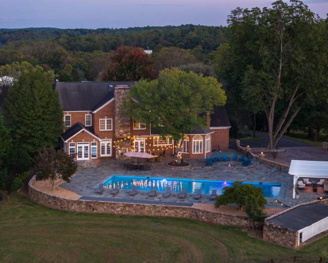 The image shows a large brick house with a swimming pool surrounded by a stone patio and landscaped yard.