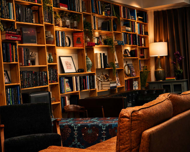 The image features a cozy interior scene with a wooden bookshelf filled with books and decorative items, flanked by comfortable seating and a lamp.