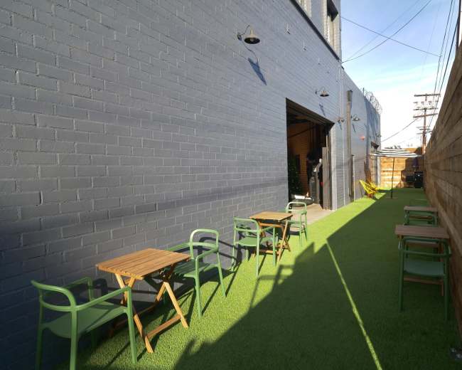 A narrow outdoor space with green artificial grass, wooden tables, and chairs along a brick wall and an open entrance to a building.