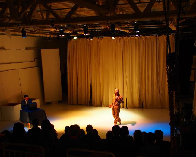 A performer stands on stage in front of a seated audience, while another individual is positioned at a table to the side, illuminated by stage lights.
