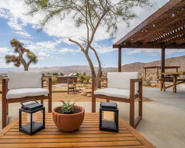 The image shows a desert patio with two white chairs, a wooden table, and a potted plant, surrounded by mountains and a cloudy sky.