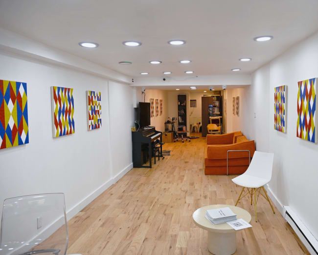 The image shows a well-lit gallery space with wooden flooring, featuring colorful geometric artwork on white walls, a piano on one side, and orange seating along with a white chair.