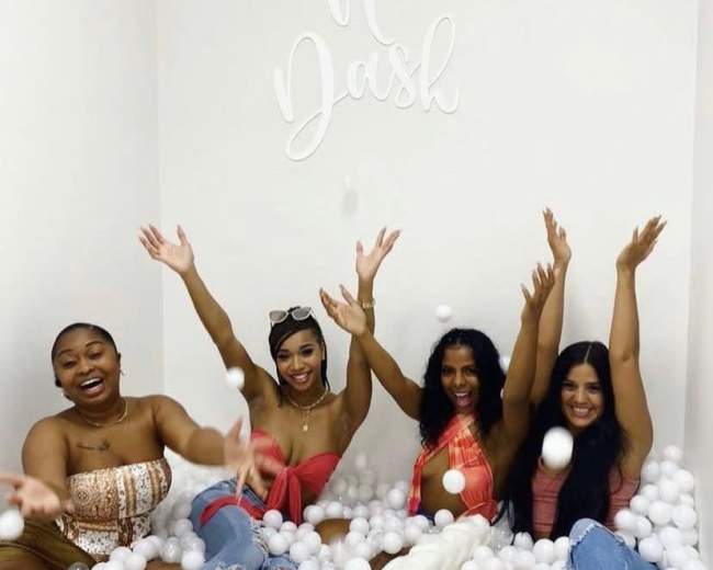 Four women are sitting in a ball pit filled with white plastic balls, wearing summer outfits and smiling as they raise their arms.