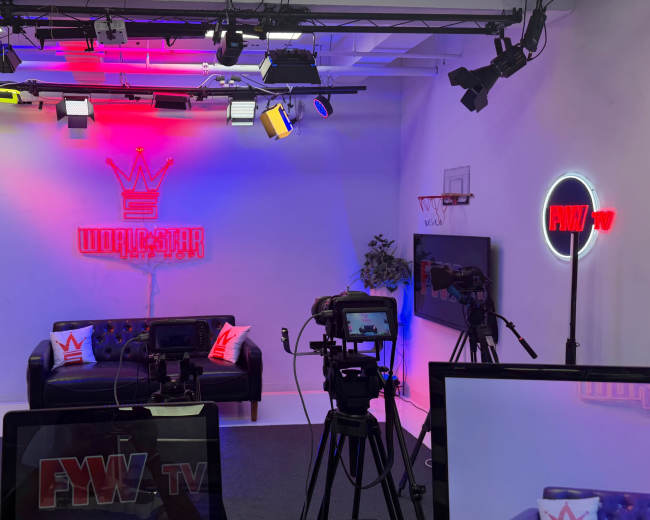 The image shows a modern studio setup with a black sofa, colorful lighting, and various cameras positioned for recording.