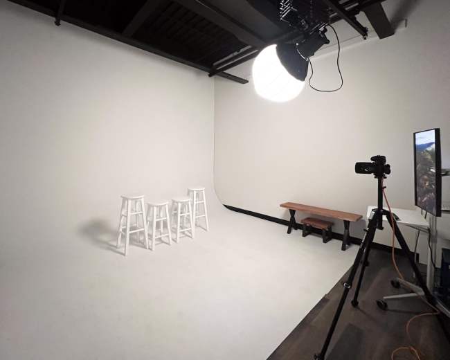A minimalist studio space featuring three white stools, a wooden bench, and a camera setup in front of a plain white backdrop.