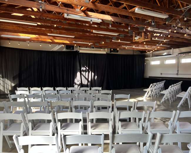 A spacious room with wooden beams and rows of white chairs facing a black curtain.