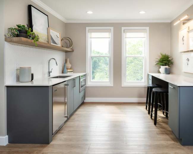 The image shows a modern kitchen with gray cabinets, white countertops, and two windows allowing natural light to enter.