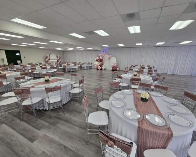 The image shows a banquet hall arranged with round tables covered in white linens, each set with plates and floral centerpieces, surrounded by white drapes and decorated with pink balloon arrangements.