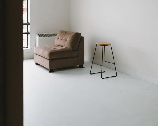 The image shows a minimalistic room featuring a brown chaise lounge and a stool positioned in front of a large window.