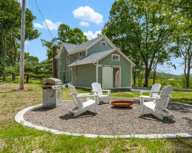 A circular seating area with white Adirondack chairs surrounds a fire pit, located in front of a two-story green house with a grill nearby and trees in the background.