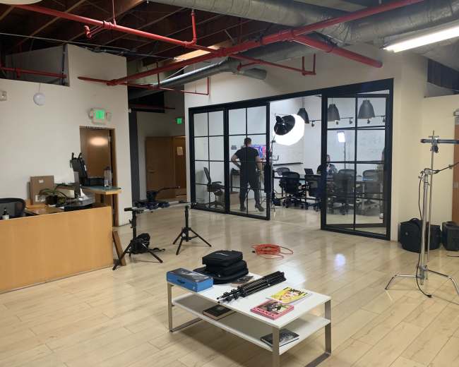 A modern office space features a reception area with a desk, a conference room in the background with people inside, and various equipment set up around the room.