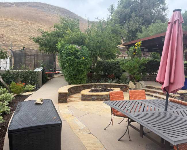 The image captures a landscaped outdoor area featuring a circular stone fire pit surrounded by greenery, a dining table with orange chairs, and a patio umbrella.