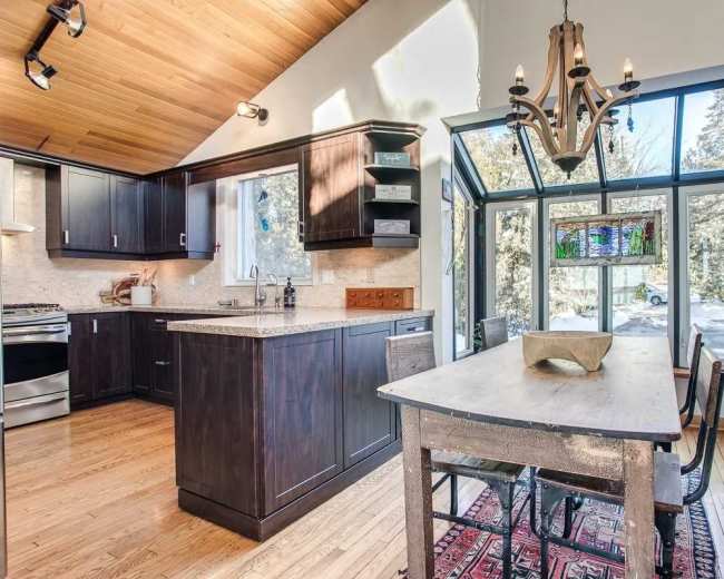 The image shows a modern kitchen featuring dark cabinetry, a granite countertop, and a dining area with a wooden table and chairs, illuminated by natural light from large windows.