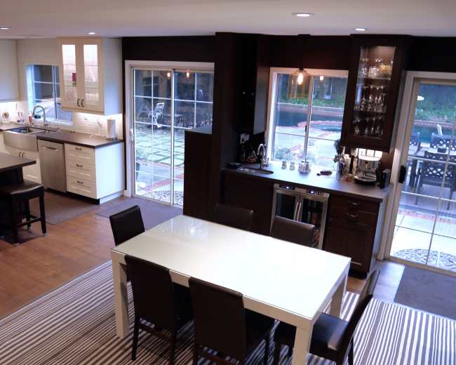 The image shows a modern kitchen and dining area with a large white table surrounded by black-chairs, complemented by a kitchen island and various appliances.