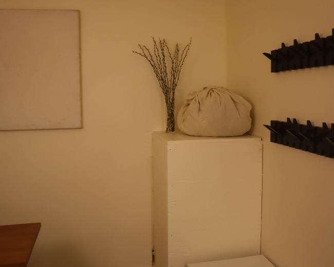 A corner of a room features a white square pedestal with a bundle of twigs on top, a fabric bag beside it, and a metal cart at the bottom.