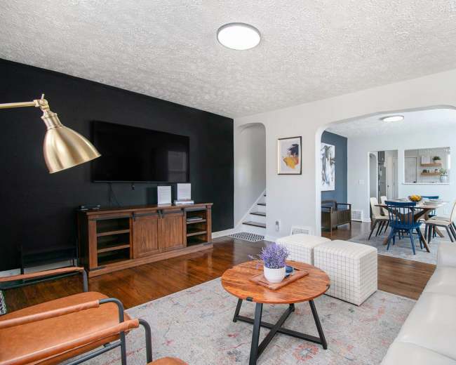 The living room features a television on a black wall, a white sofa, a wooden coffee table, and a dining area visible in the background.