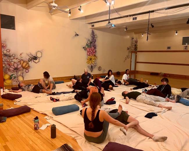 A group of people are lying on mats and cushions in a spacious room decorated with colorful murals.