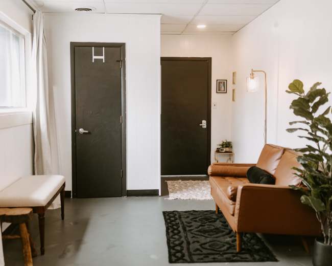 The image shows a small, minimalist waiting area featuring a brown leather sofa, a bench, a door, and a potted plant against a white wall.